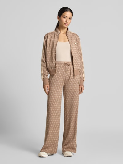 Frauenmodel trägt Sweatpants mit Logo-Muster im Allover-Look von GUESS, Beige mit Wide Leg, elastischem Bund, Tunnelzug, Ganzkörperansicht von vorne.