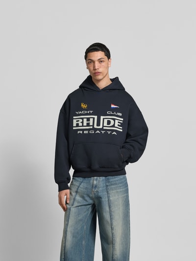 Männermodel trägt Regular Fit Hoodie mit Logo-Print von RHUDE in Dunkelblau, Reine Baumwolle, Känguru-Tasche, Elastische Abschlüsse, Ganzkörperansicht von vorne. Rückenlänge bei Größe M: 65 cm.