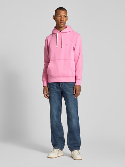 Männermodel trägt Regular Fit Hoodie aus Baumwoll-Mix von Tommy Hilfiger in Rosa mit Kängurutasche, kombiniert mit Jeans, Ganzkörperansicht von vorne.