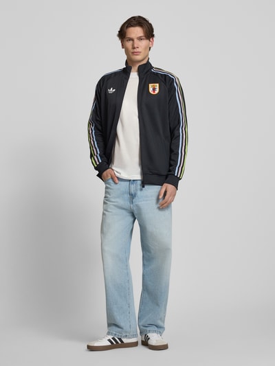 Männermodel trägt Regular Fit Trainingsjacke mit JFA-Applikation von adidas Originals, Optik: Unifarben, Stehkragen, Reißverschluss, Kontraststreifen, Logo-Stitching, Ärmelform: Langarm, Ärmelabschluss: Gerippt, Taschen: Seitliche Eingrifftaschen, Ganzkörperansicht von vorne.