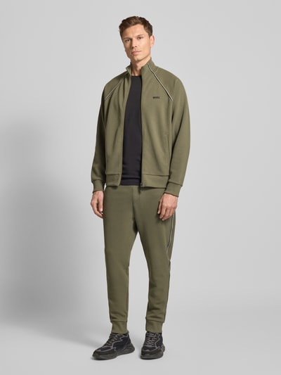 Męski model nosi Regular Fit Sweat-Anzug aus Baumwoll-Mix Modell 'TRACKSUIT SET' od BOSS Green, kolor oliwkowy, krój regular fit, jednolita struktura, detale z lamówką i paskiem z logo, materiał piqué, dwuczęściowy zestaw, zamek i kieszenie; widok całej sylwetki z przodu.