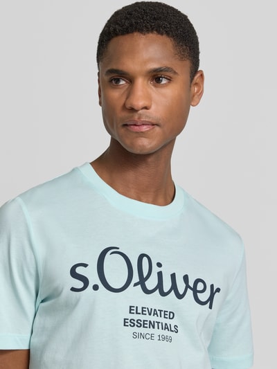Męski model nosi Regular Fit T-Shirt aus reiner Baumwolle von s.Oliver RED LABEL, efekt: jednolity kolor, krój regular fit, nadruk z logo, okrągły dekolt, krótki rękaw, dżersej, 100% bawełna, zbliżenie na górną część ciała.