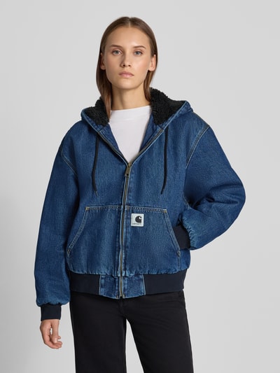 Frauenmodel trägt Jeansjacke mit Kapuze und Reißverschluss von Carhartt Work In Progress in Blau, Känguru-Tasche, Regular Fit, Ganzkörperansicht von vorne. Rückenlänge 62 cm, Schulterbreite 57 cm, Brustweite 62 cm, Ärmellänge 58 cm, reine Baumwolle, Label-Stitching.