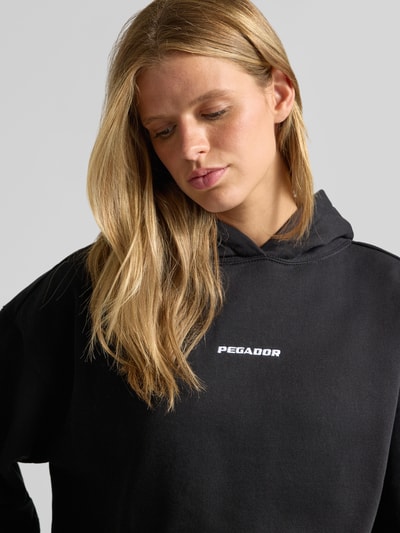 Frauenmodel trägt Relaxed Fit Hoodie aus Baumwoll-Mix Modell 'Clarita' von PEGADOR in Schwarz mit Logo-Stickerei, Nahaufnahme Oberkörper.