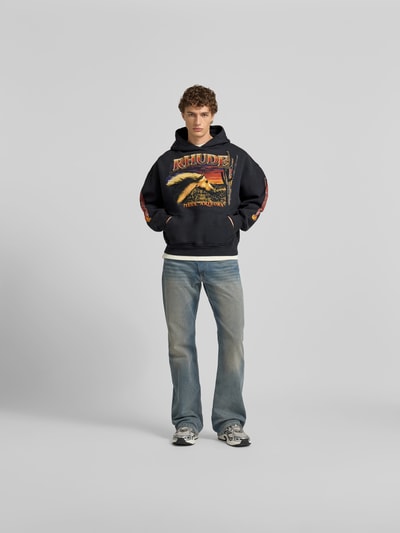 Männermodel trägt Hoodie mit Kapuze von RHUDE aus reiner Baumwolle, Label-Print, Känguru-Tasche, Lockere-Schnitt, Ganzkörperansicht von vorne.