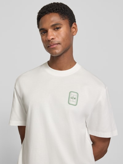 Männermodel trägt Regular Fit T-Shirt aus reiner Baumwolle Modell 'CN Tenniscourt Backprint' von Lacoste, schlichtes Weiß mit grünem Print, Herren T-Shirt, Nahaufnahme Oberkörper.