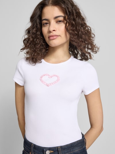 Frauenmodel trägt T-Shirt mit Rundhalsausschnitt Modell 'PREPPY HEART' von Review, weißer Baumwoll-Mix, rosa Herz, Nahaufnahme Oberkörper.