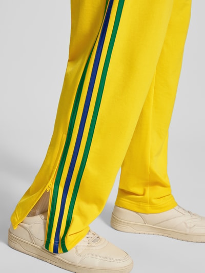 Männermodel trägt Straight Fit Sweatpants mit Logo-Stitching Modell 'Firebird' van adidas Originals in geel met blauwe en groene strepen, steekzakken met ritssluiting, elastische band, binnenbeenlengte bij maat M 40 cm, detailopname enkel.