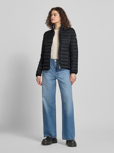 Frauenmodel trägt schwarze Steppjacke mit Logo-Stitching Modell 'FUJI' von Superdry, Gerader-Schnitt, Stehkragen, Reißverschluss, Seitliche Reißverschlusstaschen, Rückenlänge bei Größe S: 62 cm, Schulterbreite 40 cm, Brustweite 50 cm, Ärmellänge 69 cm, Ganzkörperansicht von vorne.