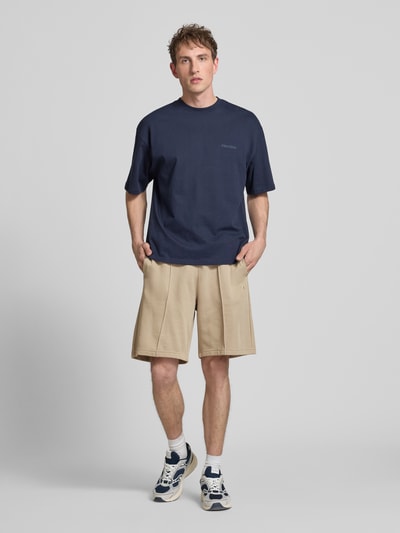 Männermodel trägt Bermudas mit elastischem Bund und Label-Stitching von CHAMPION aus Baumwoll-Mix in Beige, Ganzkörperansicht von vorne.