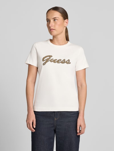 Kobiece modelka nosi T-shirt mit Logo aus Perlen od GUESS, biała bawełna, krój regular fit, okrągły dekolt, długość tyłu w rozmiarze S: 58 cm, widok całej sylwetki z przodu.