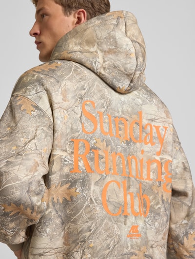 Männermodel trägt Hoodie mit Logo- und Motiv-Print von ANOTHER COTTON LAB im Camouflage-Look mit orangefarbenem Print, Ganzkörperansicht Rückansicht. Baumwoll-Mix, Loose fit, Kapuze, Känguru-Tasche, Label-Patch, Rückenlänge bei Größe M 63 cm, Ärmellänge 51 cm.