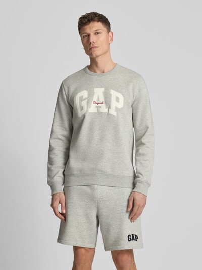 Mannenmodel draagt 'Sweatshirt met Logo en Rundhalsausschnitt' van GAP in lichtgrijs katoenmix, regular fit, logoprint, ronde hals. Ganzlichaamsaanzicht van voren.
