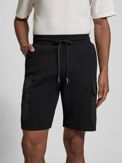 Mężczyzna nosi Regular Fit Shorts mit Cargo-Taschen od Antony Morato, mieszanka bawełny, czarne, wpuszczane i kieszenie cargo, długość wewnętrzna nogawki 23 cm, elastyczny pas z tunelem, widok na przód, zbliżenie na dolną część ciała.