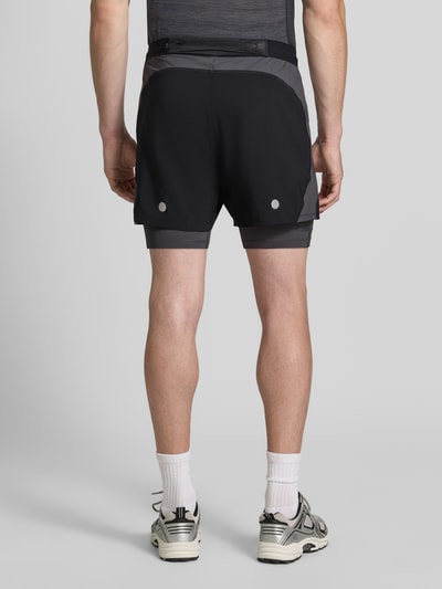 Męski model nosi Regular Fit Shorts mit integrierten Tights Model 'Road' von ASICS TIGER, czarny z szarymi detalami, widok całej sylwetki z tyłu. Szerokość w rozmiarze M: 38 cm, długość wewnętrzna nogawki w rozmiarze M: 12 cm, elastyczny pas, detal z logo.
