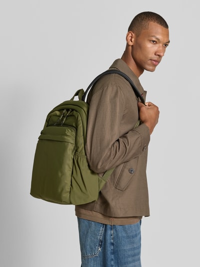 Männermodel trägt olivgrünen Rucksack mit Label-Detail Modell 'AYDEN' von HUGO, braune Jacke, strukturierte Oberfläche, Ganzkörperansicht von vorne. Herren Rucksack von HUGO.