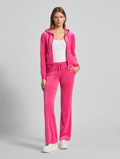 Frauenmodel trägt pinke Sweatjacke mit Label-Stitching Modell 'ROBERTSON' von Juicy Couture aus Material-Mix mit Stretch-Anteil, Regular-Fit, Kapuze mit Tunnelzug, Reißverschluss und Känguru-Tasche, Ganzkörperansicht von vorne.