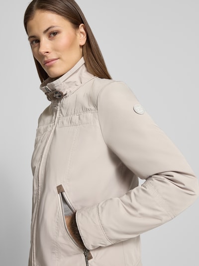Modelka kobieca ma na sobie Jacke mit Kapuze und Reißverschlusstaschen od Gil Bret, beżowa, krój regular fit, z kapturem, stojącym kołnierzem, z kieszeniami na zamek, długość tyłu w rozmiarze 36: 59 cm, szerokość ramion w rozmiarze 36: 40 cm, szerokość w biuście w rozmiarze 36: 51 cm, długość rękawa w rozmiarze 36: 63 cm, gładka faktura, widok z przodu, zbliżenie na górną część ciała.