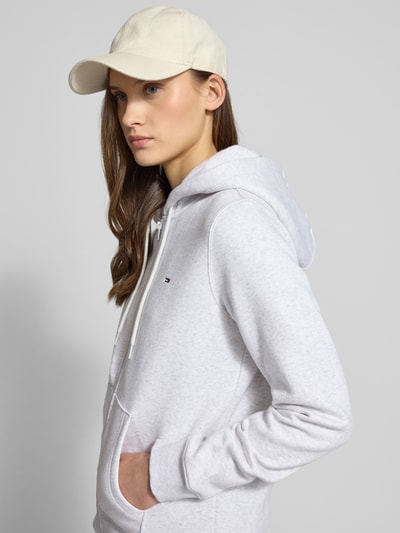 Frauenmodel trägt Regular Fit Sweatjacke aus Baumwoll-Mix von Tommy Jeans in Hellgrau, Kapuze mit Tunnelzug, Kängurutasche, Nahaufnahme Oberkörper.