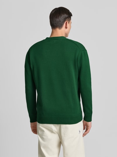 Männermodel trägt Strickjacke mit V-Ausschnitt Modell 'Tay' von Wood Wood aus grüner Baumwolle in geradem Schnitt, Ganzkörperansicht Rückansicht.