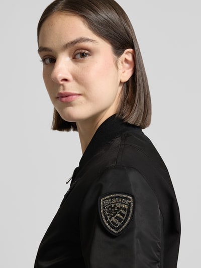 Frauenmodel trägt Bomberjacke mit Reißverschlusstaschen Modell 'LAURIE' von Blauer Usa, Schwarz, Label-Patches, Nahaufnahme Oberkörper. Damen, Loose-Fit, Stehkragen, Zweiwege-Reißverschluss, gerippte Abschlüsse, Rückenlänge Größe S: 57 cm, Schulterbreite: 44 cm, Brustweite: 43 cm, Ärmellänge: 62 cm.