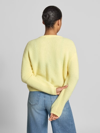 Frauenmodel trägt Relaxed Fit Strickjacke aus Woll-Mix mit Leinen-Anteil von American Vintage in Gelb, Damen Strickjacke mit gerippten Bündchen und durchgehender Knopfleiste, Langarm, kombiniert mit heller Jeans, strukturierte Oberfläche, Ganzkörperansicht Rückansicht.