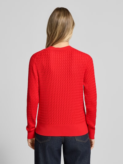 Frauenmodel trägt Slim Fit Pullover aus reiner Baumwolle von Tommy Hilfiger in Rot, Zopfmuster, Strickoptik, Logo-Detail, Ganzkörperansicht Rückansicht.