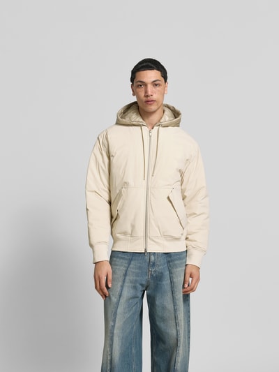 Männermodel trägt Bomberjacke mit Daunen-Feder-Füllung von Moose Knuckles in Beige, lockere Passform, Kapuze, Zweiwege-Reißverschluss, Känguru-Tasche mit Patten, glatte Textur, dazu weite Jeans, Ganzkörperansicht von vorne. Rückenlänge bei Größe M: 63 cm, Schulterbreite: 48 cm, Ärmellänge: 65 cm.