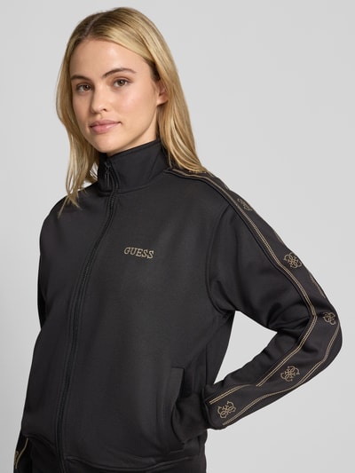 Vrouwenmodel draagt Relaxed Fit sweatjack uit elastische viscosemix van GUESS activewear, opstaande kraag, Strass-steentjes, Sierrand, elastische afwerkingen, labeldetail, zwarte kleur, gladde stof, geheelopname bovenlichaam van voren.