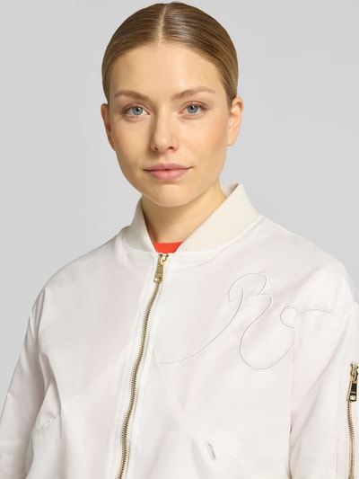 Kobiece modelka nosi Bomberjacke mit Zweiwege-Reißverschluss od Original Bombers, kolor kremowy, krój regular fit, stójka, tekstura z przeszyciami, widoczny dwukierunkowy zamek, detal rękawa z zamkiem, szerokość w rozmiarze S: 56 cm, długość tyłu w rozmiarze S: 55 cm, szerokość ramion w rozmiarze S: 50 cm, długość rękawa w rozmiarze S: 64 cm, zbliżenie na górną część ciała z przodu