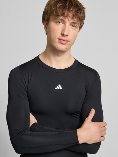 Mannenmodel draagt Longsleeve mit Label-Print van adidas Training in zwart, slim fit, geribde ronde hals, materiaal-mix met stretch, Nahaufnahme bovenlichaam.
