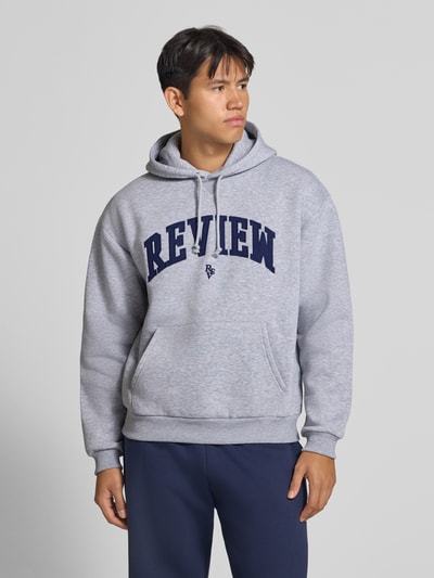 Männermodel trägt Hoodie mit Label-Stitching von REVIEW in hellgrau mit blauer Schrift, lockere Passform, Ganzkörperansicht von vorne. Baumwollmischung, Kapuze mit Tunnelzug, Känguru-Tasche, gerippte Abschlüsse, Rückenlänge 64 cm, Ärmellänge 75 cm.