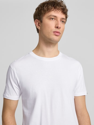 Männermodel trägt T-Shirt aus Baumwolle von Alpha Industries in Weiß mit Label-Details, Gerader-Schnitt, Rundhalsausschnitt, Nahaufnahme Oberkörper. Herren T-Shirt, Rückenlänge bei Größe M: 68 cm, Reine Baumwolle.