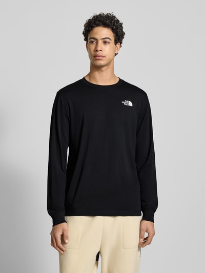 Męski model nosi Regular Fit Longsleeve mit Logo-Print od The North Face, czarny bawełniany longsleeve, krój regular fit, nadruk logo, okrągły dekolt, długie rękawy, elastyczne mankiety, długość pleców w rozmiarze M: 69 cm, zdjęcie całej sylwetki z przodu.
