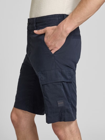 Männermodel trägt dunkelblaue Relaxed Fit Cargoshorts aus Baumwoll-Mix Modell 'SISLA' von BOSS Orange mit Label Patch und Cargotaschen, Nahaufnahme Oberschenkel.