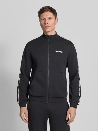Męski model nosi Sweatjacke mit Stehkragen und Zweiwege-Reißverschluss od EA7 Emporio Armani, krój regular fit, czarny kolor, widok całej sylwetki z przodu.