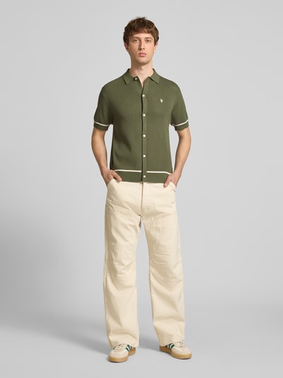 Männermodel trägt Slim Fit Strick-Poloshirt mit Label-Stitching von REVIEW, oliv mit Kontraststreifen, Knopfleiste, Ganzkörperansicht von vorne.