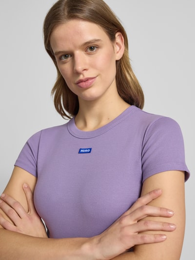 Modelka nosi damski t-shirt Slim Fit T-Shirt aus Baumwoll-Mix Modell 'Baby Tee' od Hugo Blue, jednokolorowy, mieszanka bawełny, detal z logo, okrągły dekolt, krótki rękaw, wykończenie: dżersej, kolor fioletowy, efekt: jednolity kolor, krój slim fit, skrócony krój, detal z logo, ujęcie: zbliżenie górnej części ciała.