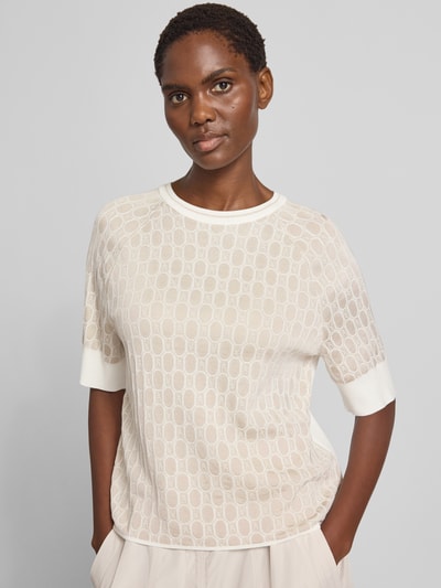 Frauenmodel trägt Relaxed Fit Bluse aus Viskose-Mix mit Allover-Logo-Detail von Marc Cain, cremefarbene Strick-Optik, Halbarm, Rundhalsausschnitt, dezentes Logo-Muster, Rückenlänge 54 cm, Ärmellänge 32 cm, Nahaufnahme Oberkörper.