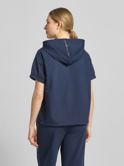 Modelka nosi T-Shirt aus Baumwoll-Mix mit Kapuze Modell 'SHARON3' od FIRE + ICE w kolorze granatowym, luźny fason, bawełna z elastanem, krótki rękaw, elastyczne mankiety, kaptur i tunel w pasie, detal z logo, widok całej sylwetki od tyłu.