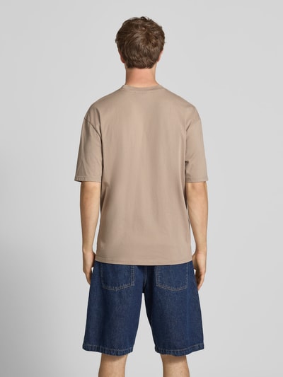 Männermodel trägt Oversized T-Shirt mit Label-Stitching von Pegador in Beige, kombiniert mit dunkelblauer Jeansshorts, Ganzkörperansicht Rückansicht. Oversized Herren T-Shirt aus Baumwoll-Mix mit Rundhalsausschnitt, Rückenlänge Größe M 70 cm.