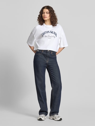 Frauenmodel trägt weißes Cropped T-Shirt mit Logo-Print von Calvin Klein Jeans aus reiner Baumwolle, kombiniert mit dunkelblauer Jeans; Ganzkörperansicht von vorne.