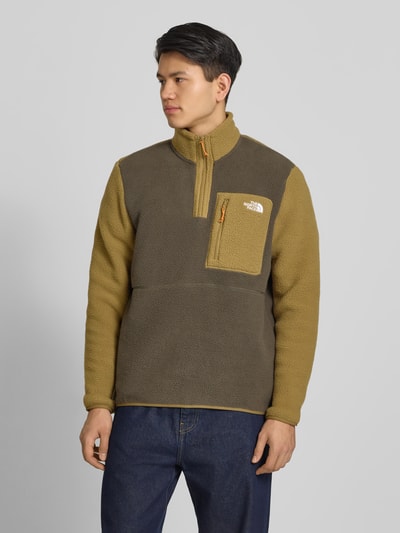 Męski model nosi Fleecepullover mit Brusttasche Modell 'YUMIORI' von The North Face, brązowo-oliwkowy, krój regular fit, stójka, zamek błyskawiczny, brustasza, wyhaftowane logo, długość tyłu w rozmiarze M: 70 cm, długość rękawów: 67 cm, ganzkörperansicht von vorne.
