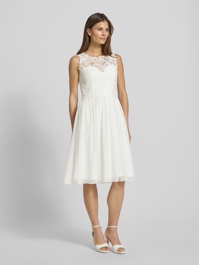 Kobieta modelka nosi Brautkleid mit floraler Spitze od Mascara, taliowany krój, szerokie ramiączka, dekolt w kształcie serca, półprzezroczysty materiał, halka pod sukienkę, zapinanie na haftkę i wszyty zamek błyskawiczny, długość tyłu 107 cm, szerokość klatki piersiowej 40 cm, kolor biały, faktura koronki, kwiatowy wzór, widok całej sylwetki z przodu.