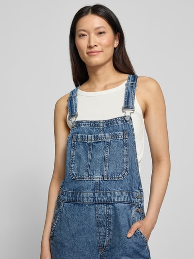 Vrouwenmodel draagt Barrel Fit Denim-tuinbroek met verstelbare bandjes van GAP, puur katoen, borstzak, denimstructuur, nahaufnahme bovenlichaam.