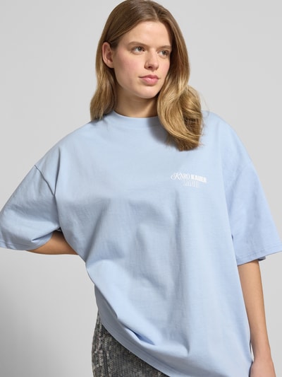 Frauenmodel trägt Oversized T-Shirt mit rückseitigem Statement-Print von Karo Kauer aus Reine Baumwolle, Oversized-Fit, Rundhalsausschnitt, Label-Print, helle Blautöne, weiche Textur, überschnittene Schultern, Rückenlänge 71 cm bei Größe S, Nahaufnahme Oberkörper.