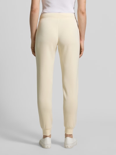 Frauenmodel trägt cremefarbene Sweatpants mit Label-Detail Modell 'OLYMPE' von Guess Activewear, körpernaher Schnitt, Ganzkörperansicht Rückansicht.