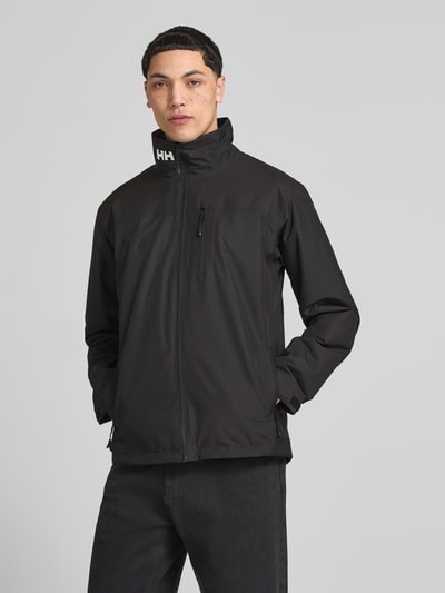 Męski model nosi Midlayer Jacke mit Stehkragen Modell 'CREW' od Helly Hansen, czarna, prosty krój, zamek błyskawiczny, nadruk z logo, brustasza zapinana na zamek błyskawiczny, naszywka z logo, boczne kieszenie zapinane na zamek błyskawiczny, długość tyłu w rozmiarze M: 74 cm, szerokość ramion w rozmiarze M: 49 cm, szerokość w rozmiarze M: 52 cm, długość rękawów w rozmiarze M: 68 cm, miękka tekstura, całkowity widok z przodu