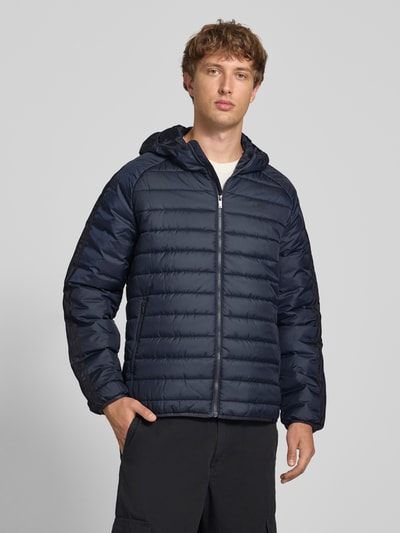 Männermodel trägt Regular Fit Steppjacke mit Reißverschluss von adidas Originals in Dunkelblau, glatte Textur, Ganzkörperansicht von vorne.