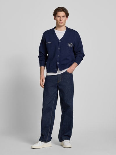 Männermodel trägt T-Shirt aus Baumwoll-Leinen-Mix mit Brusttasche von Superdry in Weiß, kombiniert mit Cardigan, Regular-Fit, Ganzkörperansicht von vorne. Herren T-Shirt, Baumwoll-Leinen-Mix, Regular-Fit, Rundhalsausschnitt, Brusttasche, Rückenlänge bei Größe M: 66 cm.